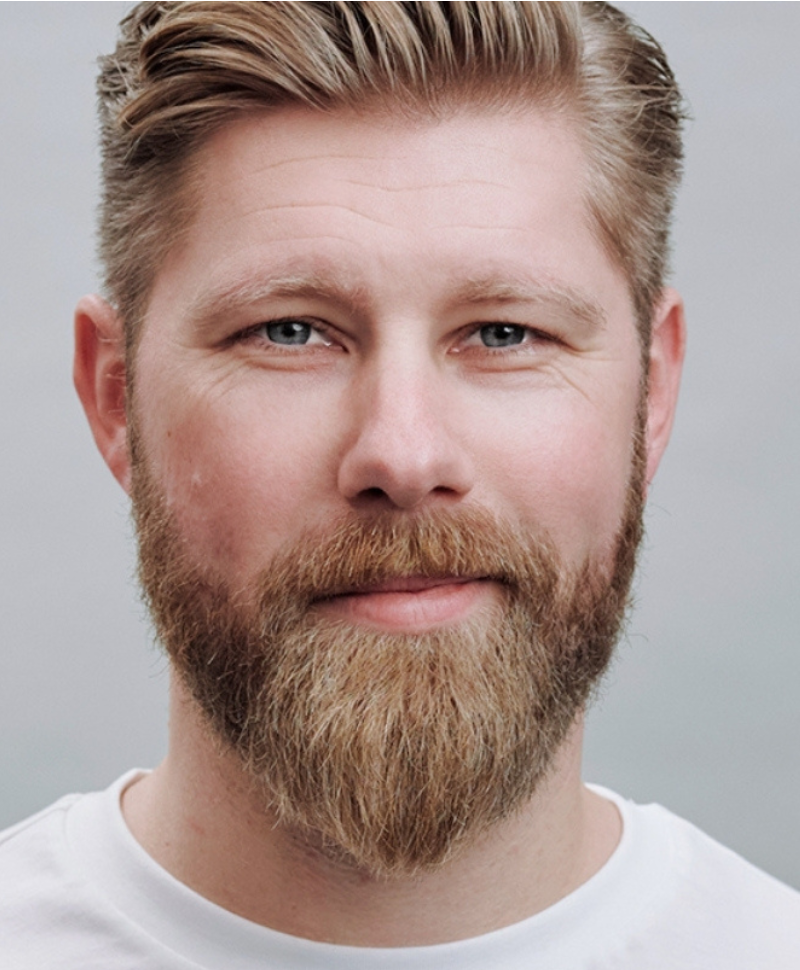 Portrait eines Mannes mit blondem Vollbart und weißem T-Shirt vor hellem Hintergrund – neutral und freundlich.