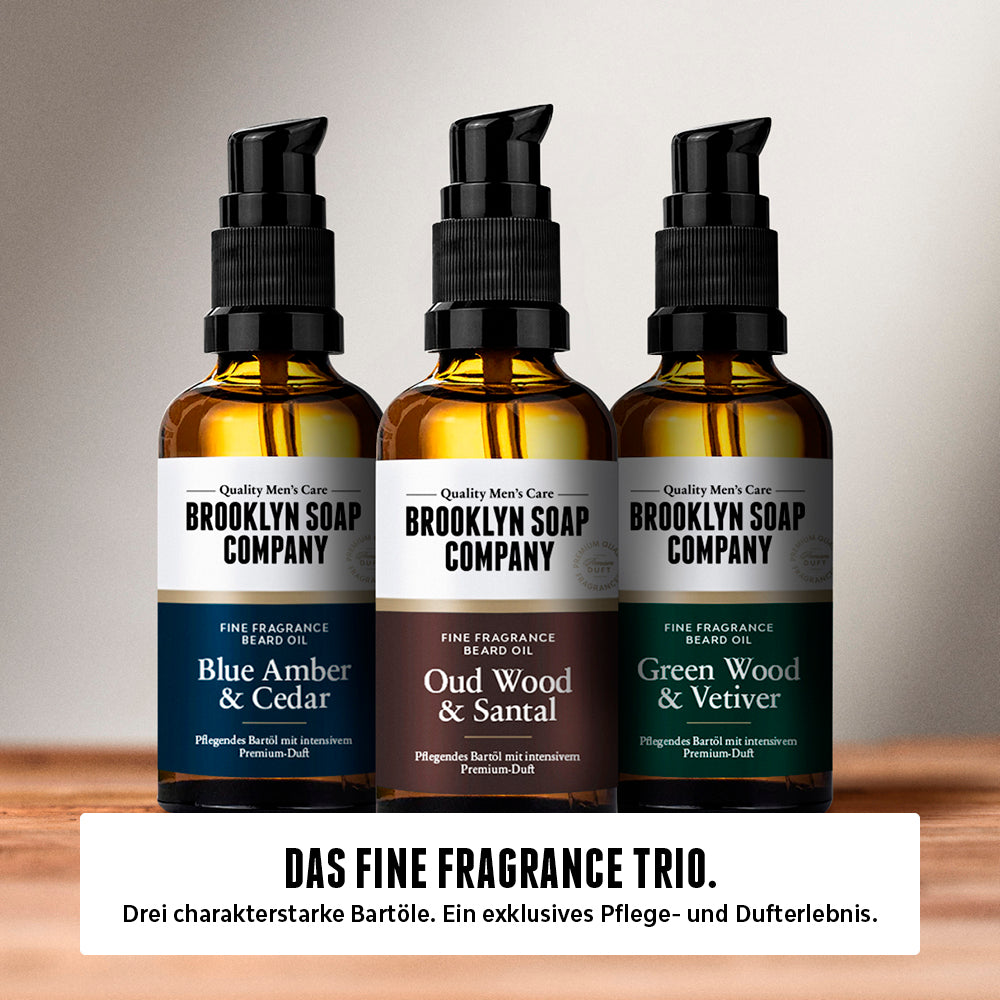 Drei Bartöle der Brooklyn Soap Company – Blue Amber & Cedar, Oud Wood & Santal sowie Green Wood & Vetiver – in bernsteinfarbenen Glasflaschen mit Pumpspender, auf einer Holzoberfläche vor einem sanft verlaufenden hellgrauen Hintergrund