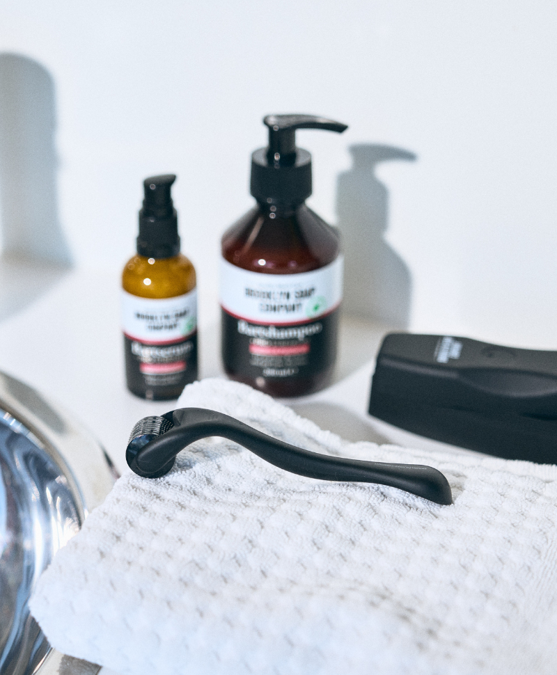 Bartroller auf Handtuch mit Bartshampoo, Bartserum und Pflegeprodukten von Brooklyn Soap Company.
